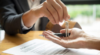 Person handing over a key to another person with a pen and document on a table.