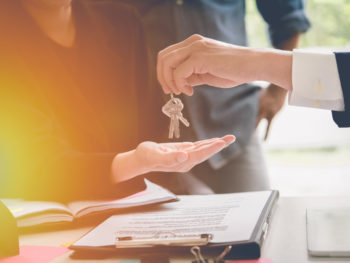 Person handing over keys to another person with a laptop and documents on a table.