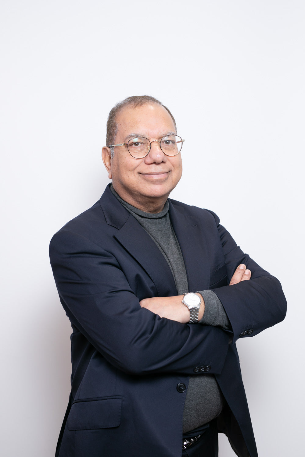 Man wearing a dark blue blazer and gray sweater with arms crossed against a white background