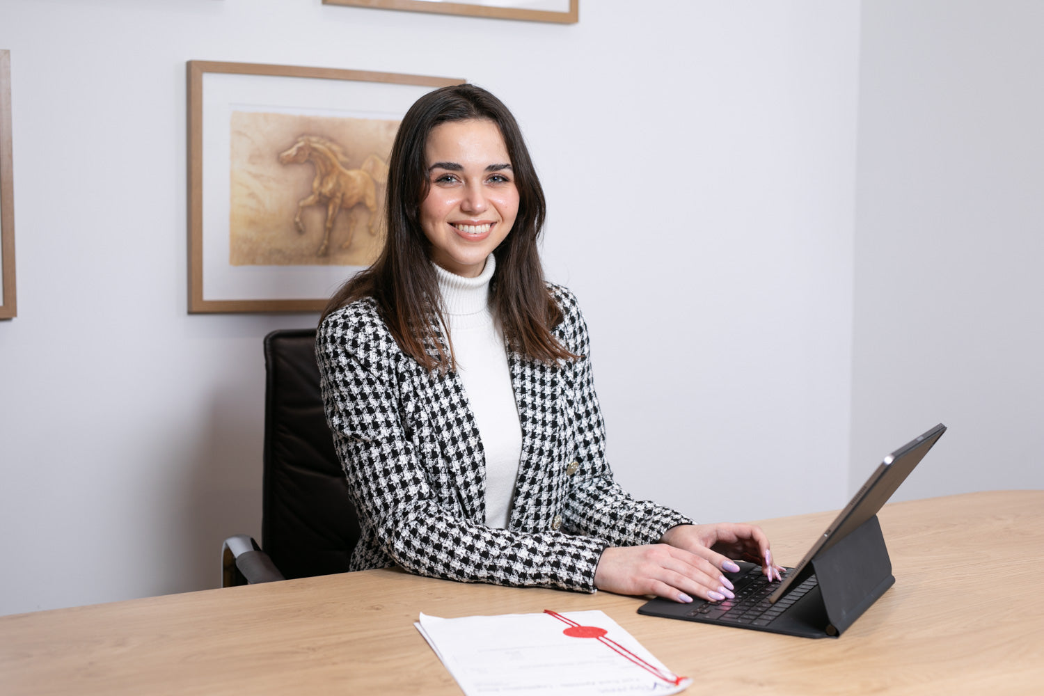 Woman sitting at a desk with a tablet, smiling in an office setting.