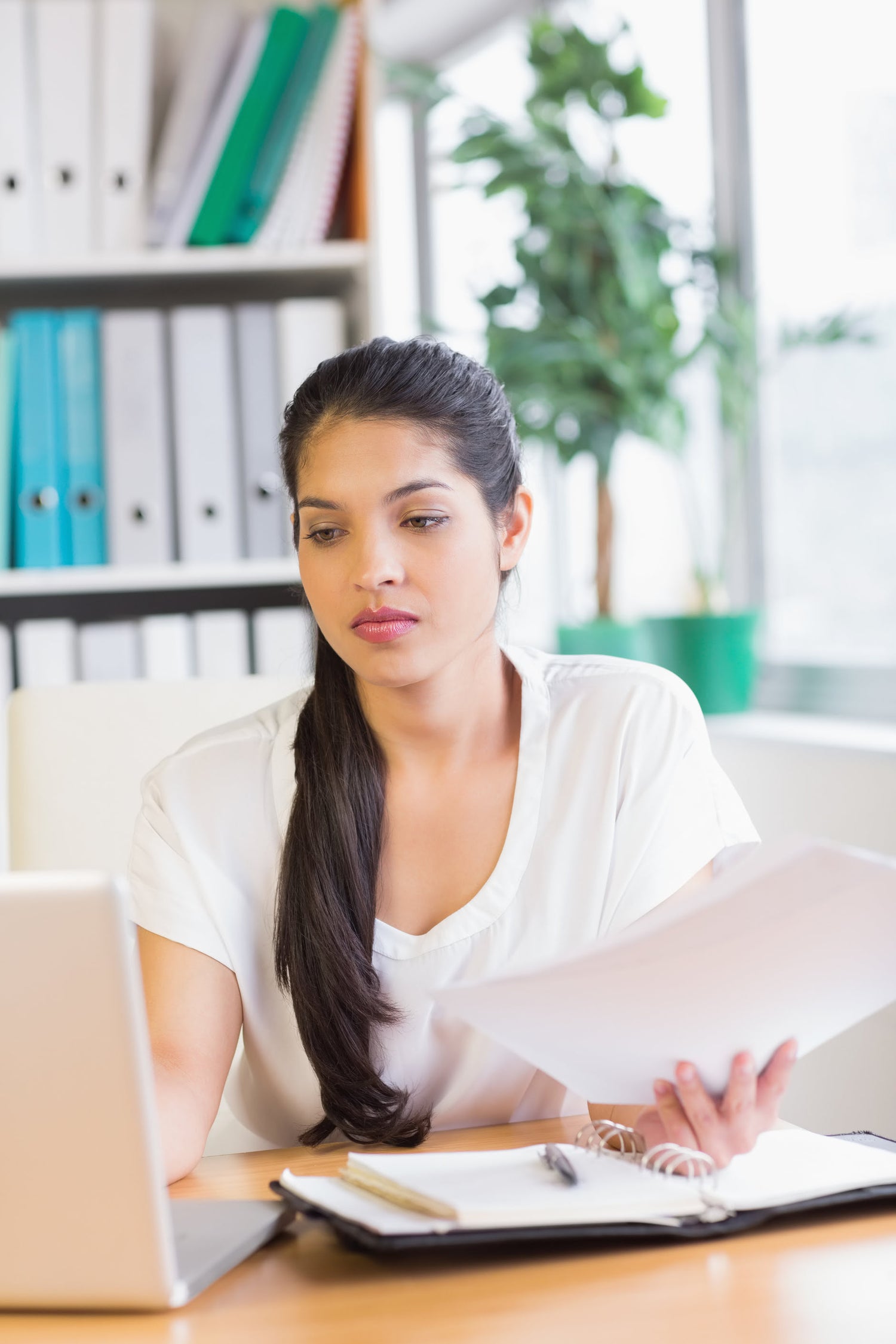 Woman sitting at a desk with a laptop and documents, looking at the camera.