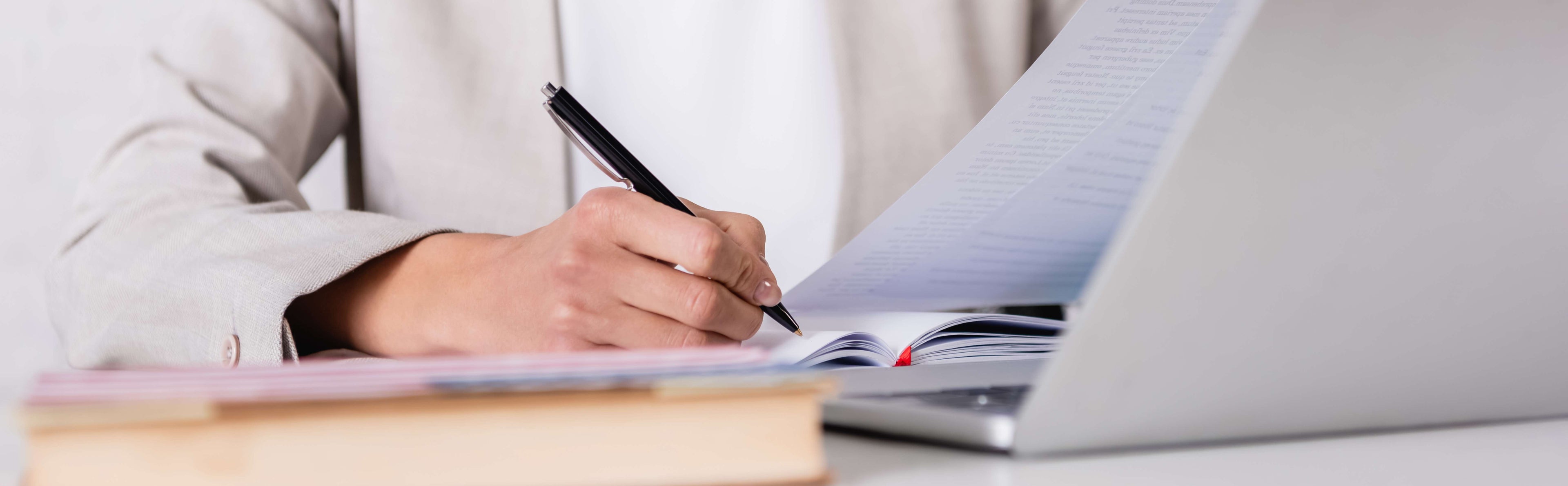 Person taking notes with a pen and notebook next to a laptop on a desk.