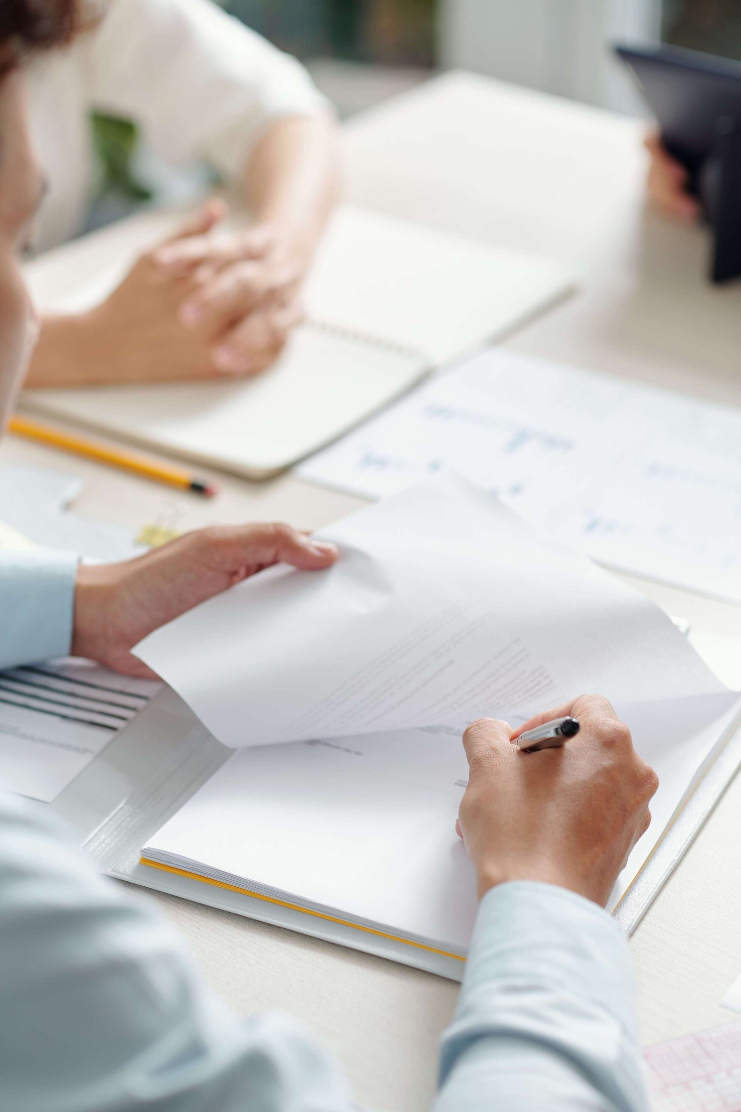 Person writing in a notebook with another person in the background.