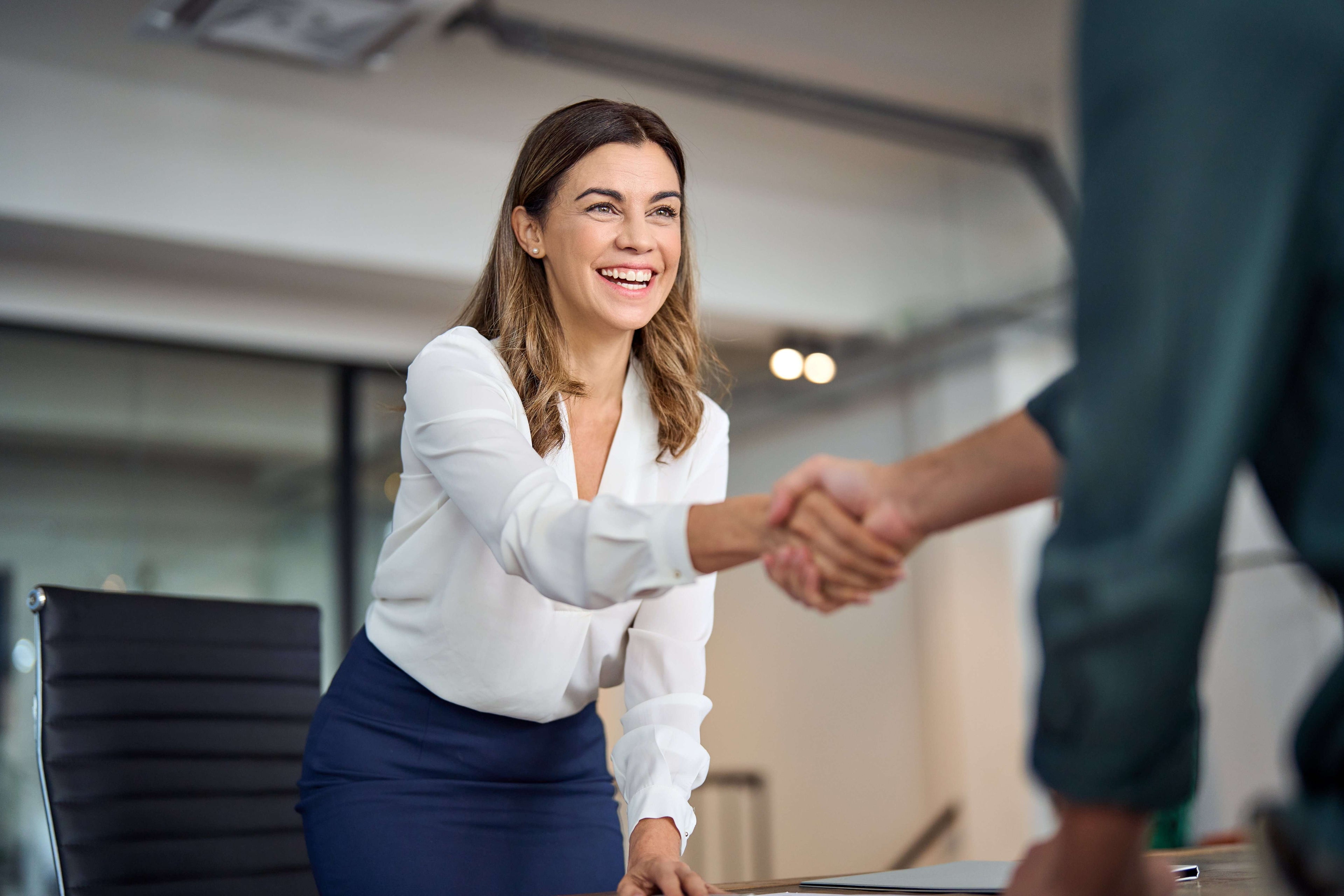 Woman in a white blouse and blue skirt shaking hands with a man in an office setting.